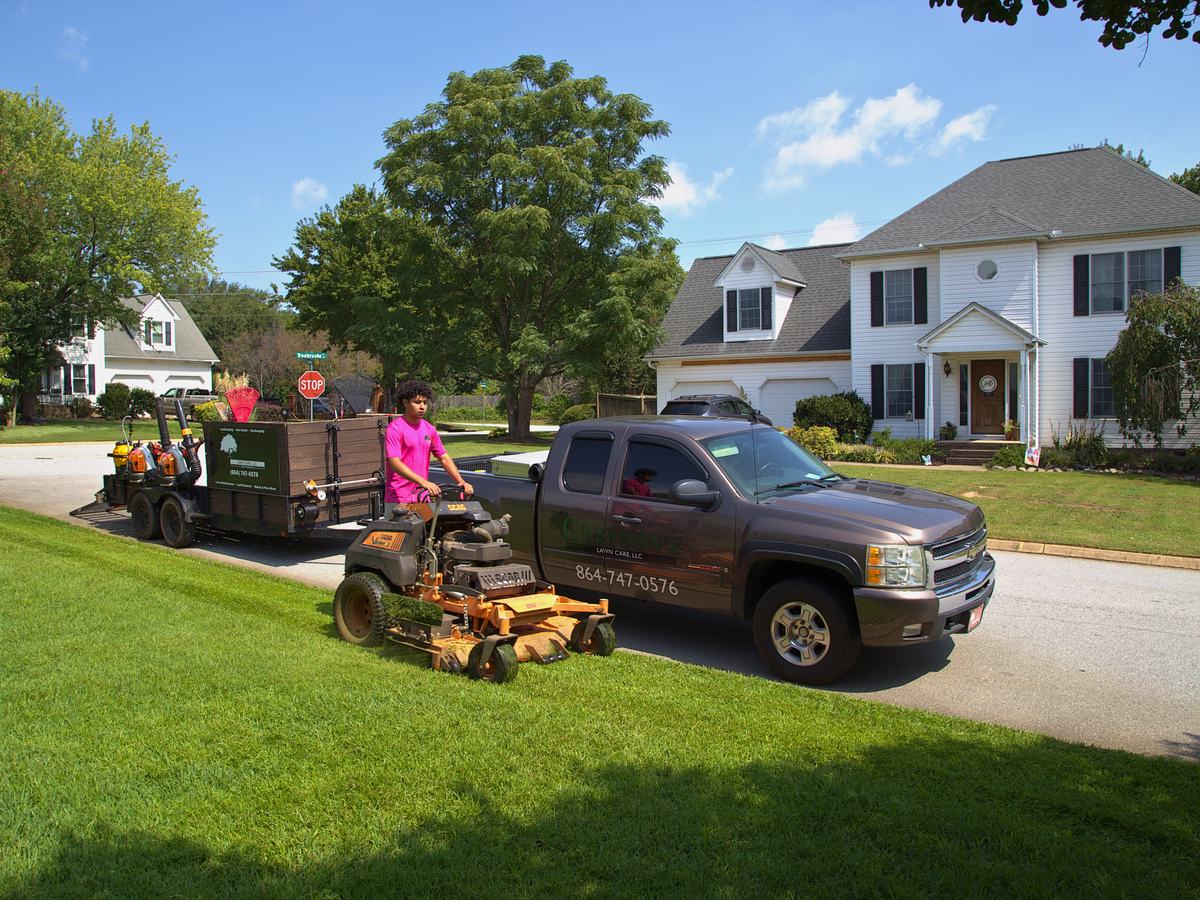 Mowing a sunny front lawn beside the company truck and trailer.